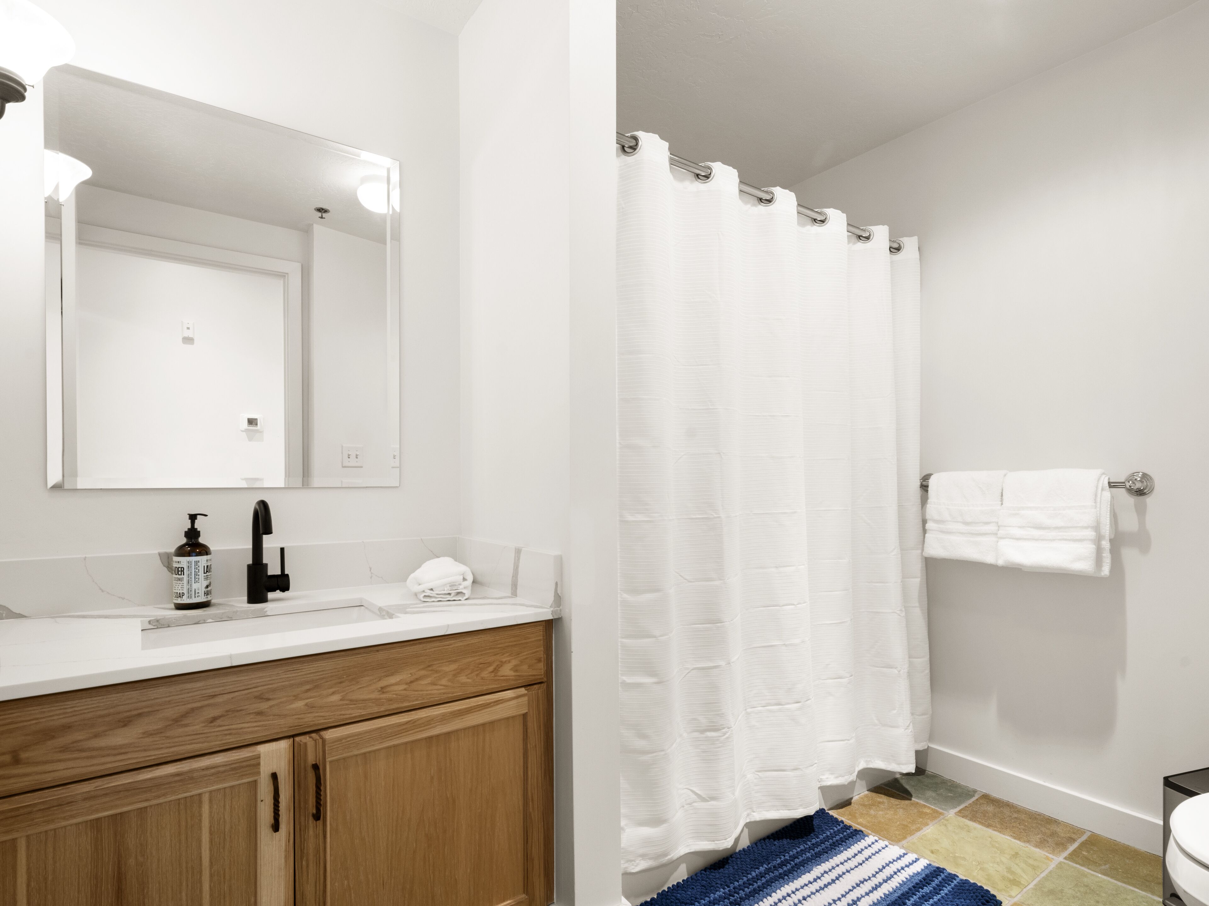 Une salle de bain avec un lavabo blanc, des meubles en bois, un miroir, un rideau de douche blanc et un tapis à rayures bleues.