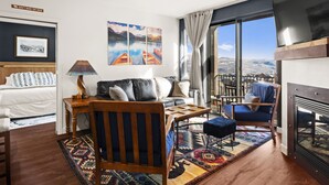 Une chambre d’hôtel moderne équipée d’un lit, d’un canapé, d’une table à manger, d’une télévision et d’une grande fenêtre donnant sur les montagnes.