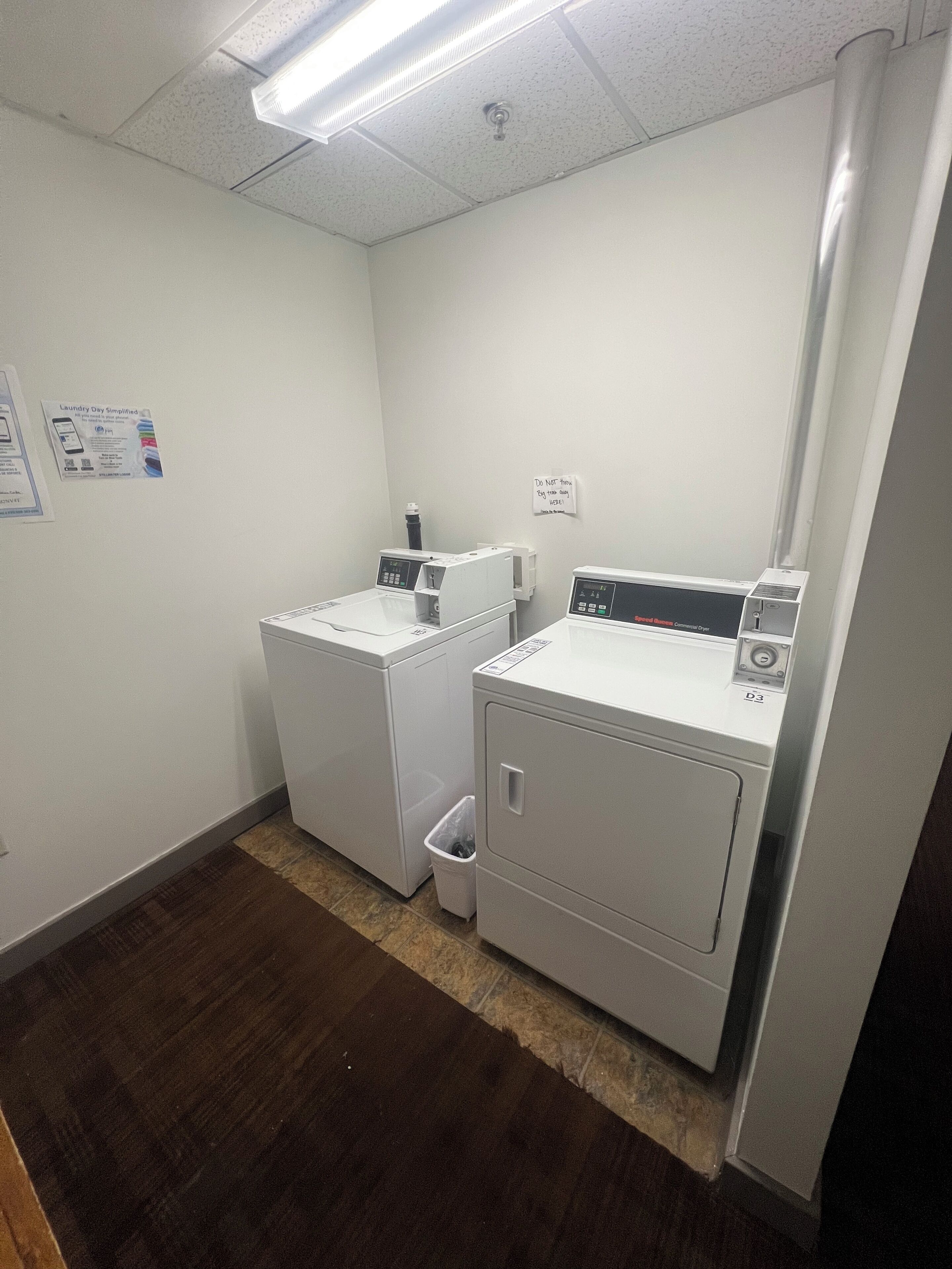 A laundry room with two white front-loading washers and dryers, a trash can, and a sign on the wall.