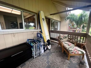 Un porche cubierto con una tabla de surf apoyada en la pared, un banco y una silla con cojín floral.