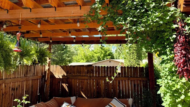 A covered outdoor seating area with a wooden pergola, cushioned seating, and hanging plants. - Casa del Sol (Taos)