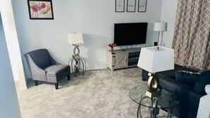 Una sala de estar con un sofá gris, un sillón negro, una mesa de centro de vidrio, un televisor y un aparador.