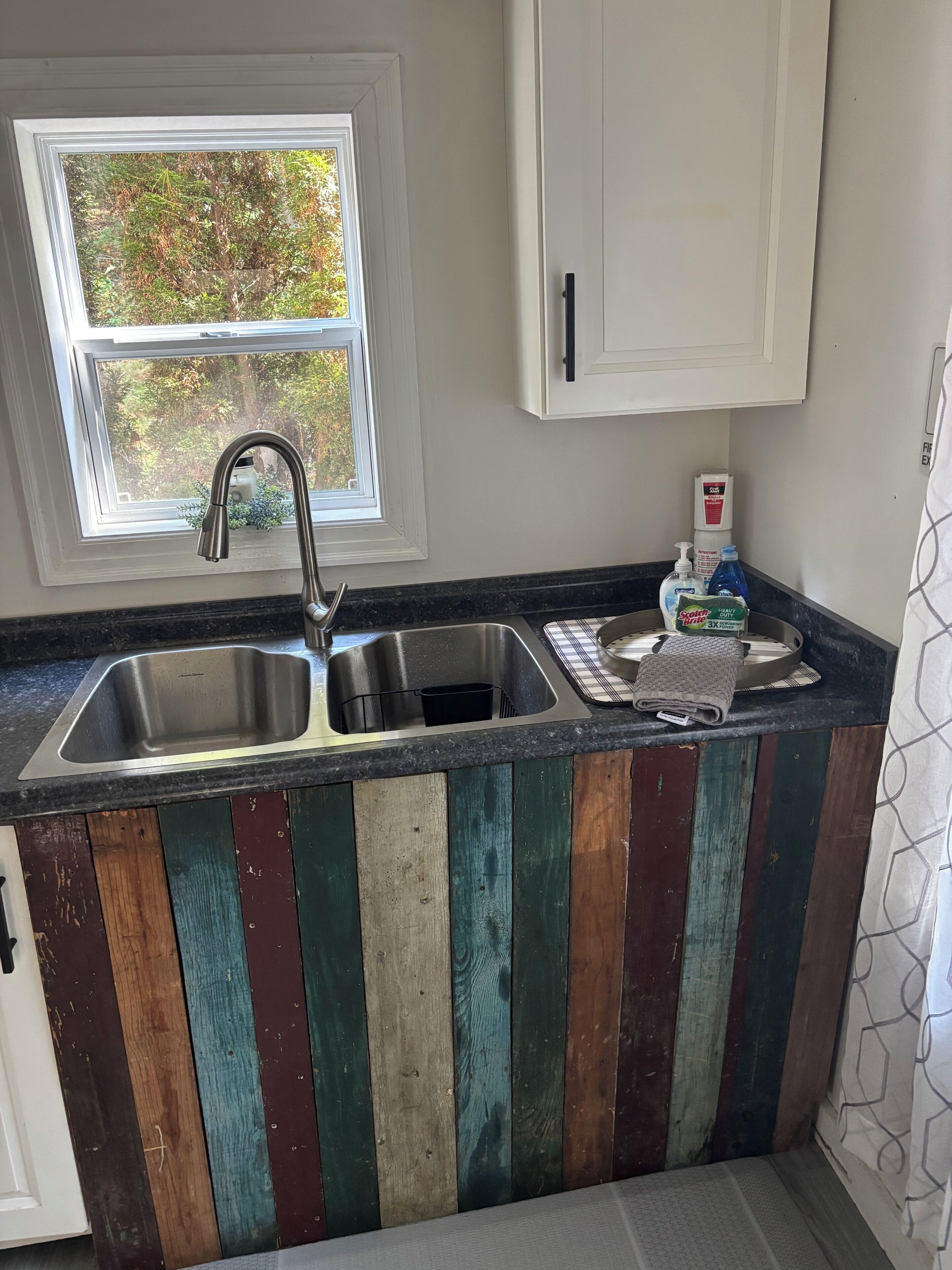 A kitchen with a double sink, wooden panel backsplash, a window with a view of trees, and a cabinet above the sink.