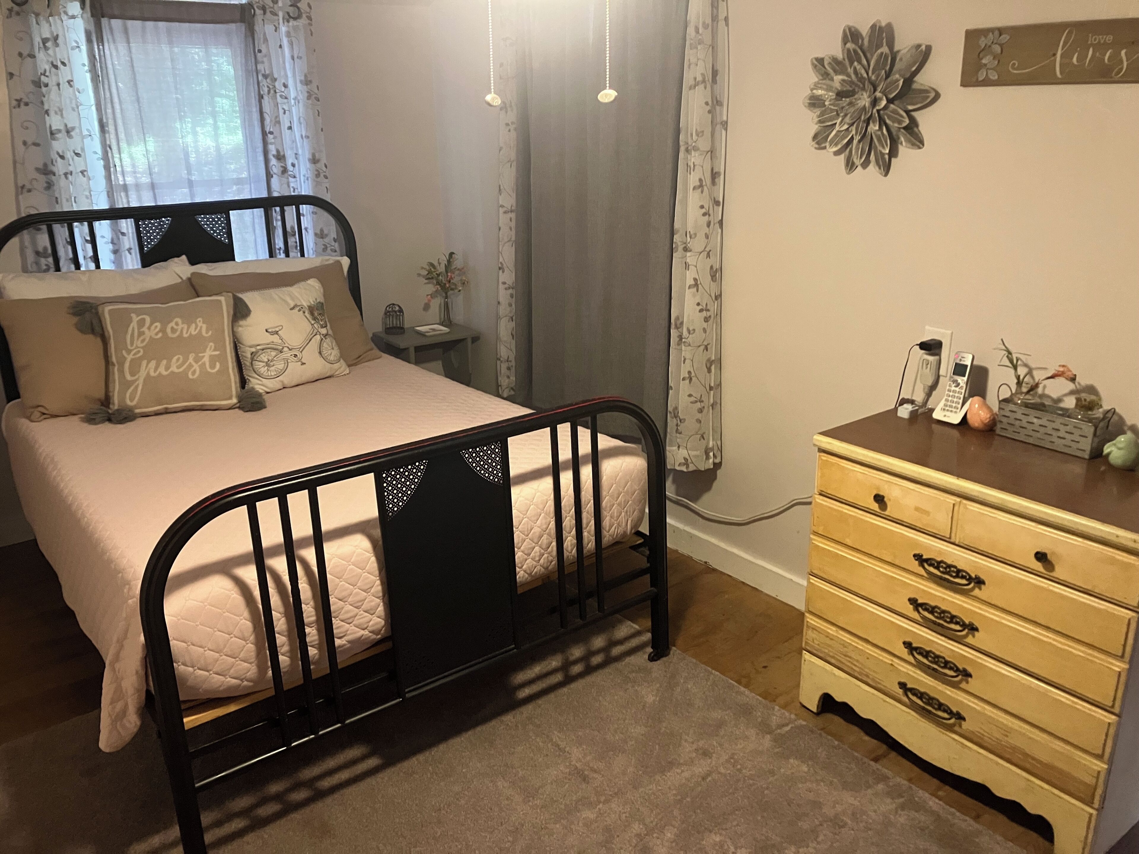 Ein Schlafzimmer mit Bett, Kommode, Nachttisch und einem Fenster mit Vorhängen.