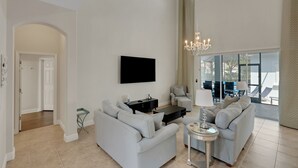 A spacious living room with a large sofa, a coffee table, and a TV mounted on the wall. There is a chandelier hanging from the ceiling and a view of the outdoors through large windows. - GUARDIANS and Mickey Themed! Stunning! Reunion Resort! (Reunion)