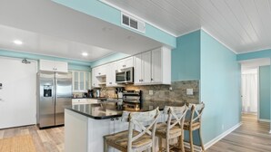 A modern kitchen with white cabinets, a stainless steel refrigerator, a microwave, and a granite countertop. There are wooden chairs and a small island with a sink.