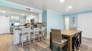 A modern kitchen with white cabinets, a stainless steel refrigerator, and a dining area with a wooden table and wicker chairs.