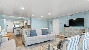 A modern living room with a sofa, striped armchairs, a flat-screen TV, and a kitchen in the background.