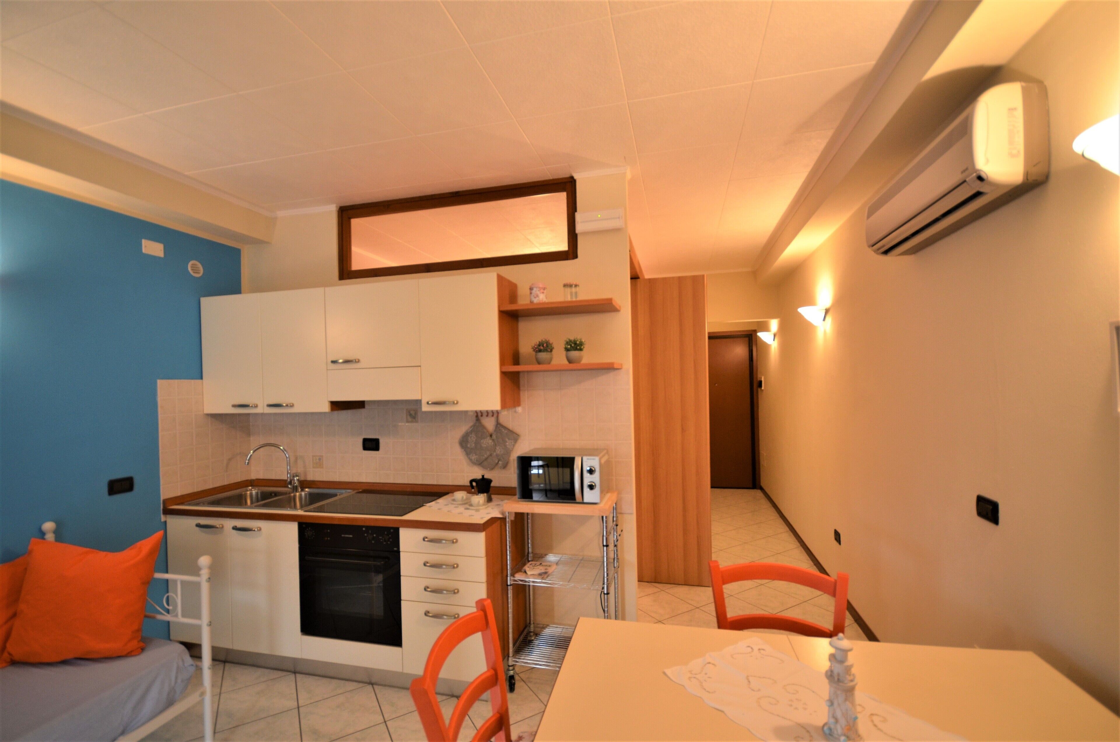 A compact kitchen with white cabinets, a microwave, and a sink. There is a small dining table with chairs and a bed with an orange pillow.