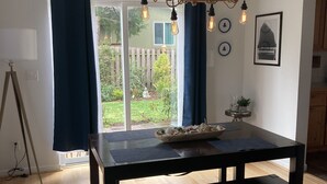 A dining area with a dark wooden table, bench, and blue rug. A chandelier hangs above, and a window with curtains is visible.