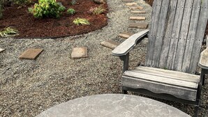 A patio area with a fire pit, a wooden Adirondack chair, and a gravel path leading to a wooden fence.