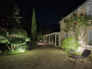 A paved pathway leading to a building with a glass door entrance, surrounded by well-maintained greenery and a bench.