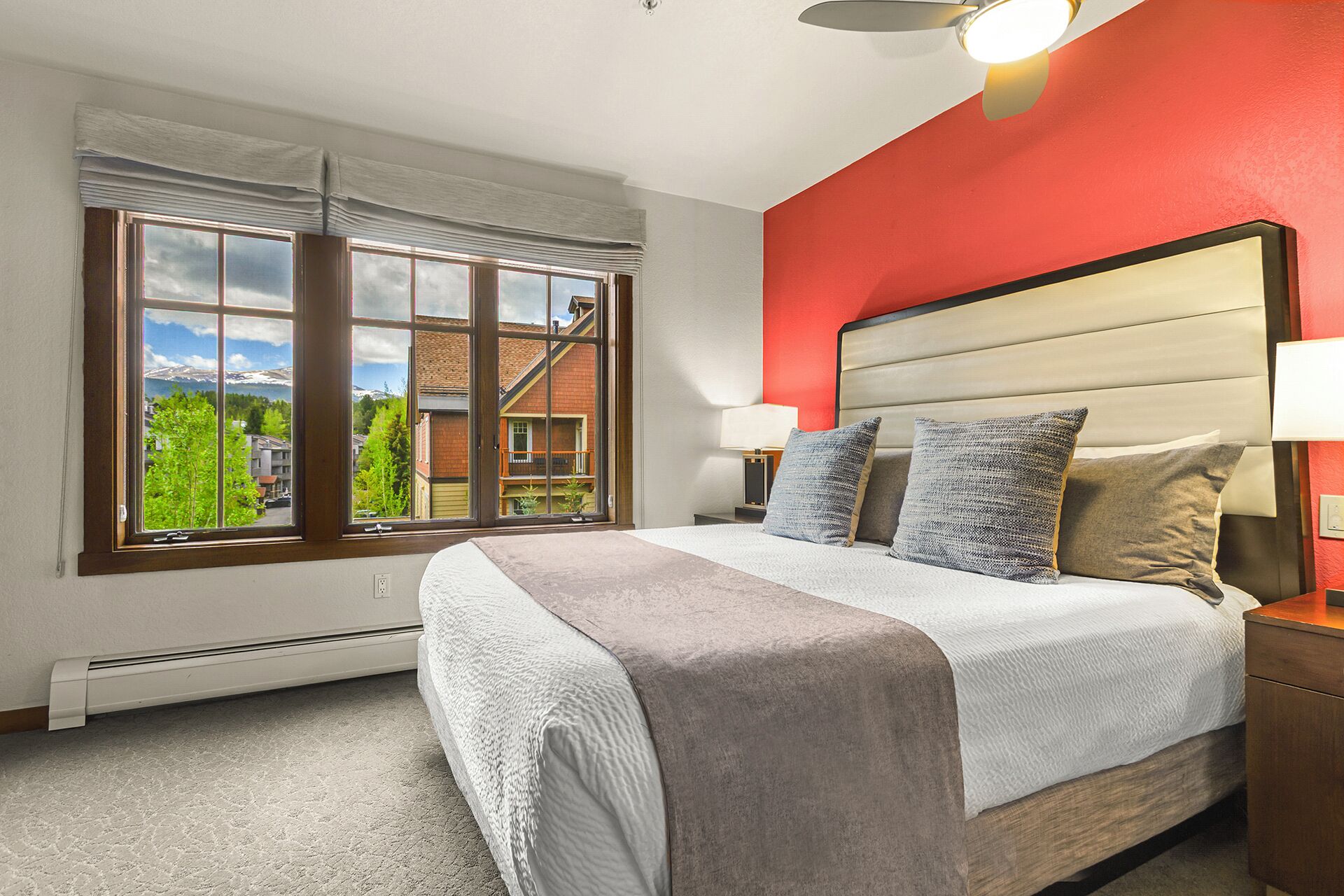 Ein Schlafzimmer mit einem großen Bett, zwei Nachttischen, einem Fenster mit Blick auf Gebäude und Bäume sowie einer roten Akzentwand.