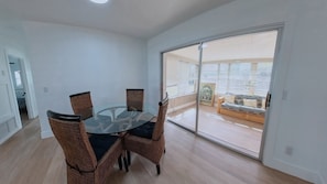 Un'area pranzo con tavolo in vetro e sedie in vimini, una porta scorrevole in vetro che dà su un balcone e un quadro appeso al muro.