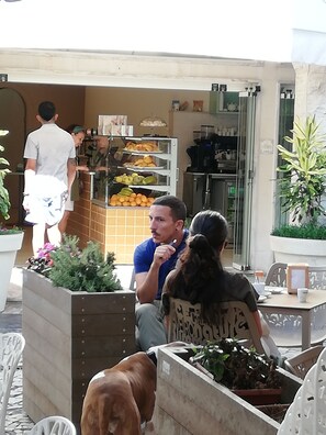 A man sitting at an outdoor cafe with a woman and a dog. There are potted plants and a food display in the background.