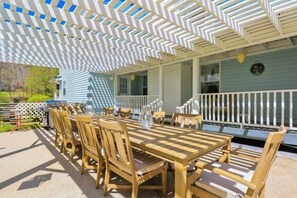 An outdoor patio with a wooden table and chairs, a white lattice ceiling, and a blue house in the background. - Spacious Murphy Home w/ Game Room + Fire Pit! (Murphy)
