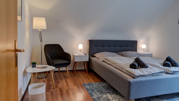 A bedroom with a bed, bedside tables, a chair, a small table, and a lamp. - Gemütliche Ferienwohnung im Grünen (Alsfeld)