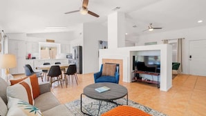 Un'ampia zona giorno con divano, tavolino e TV sullo sfondo. È presente una zona pranzo con sedie e tavolo, e una cucina con pensili ed elettrodomestici.