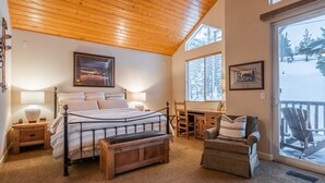 A bedroom with a bed, bedside tables, a bench, a chair, and a desk. There is a window with a view of trees and a deck outside.
