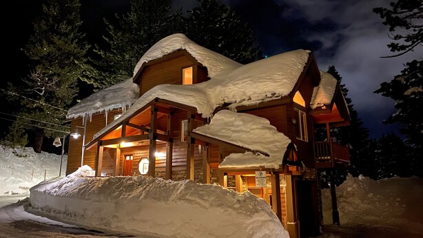 A wooden cabin with a sloped roof covered in snow, a lit interior, and a 'No Parking' sign in the foreground.