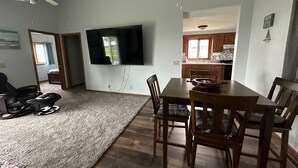 Un salon doté d’un téléviseur à écran plat fixé au mur, d’une table à manger avec des chaises, d’un ventilateur de plafond et d’un coin cuisine visible à travers une porte ouverte.