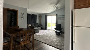 Un vaste espace de vie doté d’un ventilateur de plafond, d’une table à manger avec des chaises, d’un canapé et d’une grande fenêtre agrémentée de rideaux.