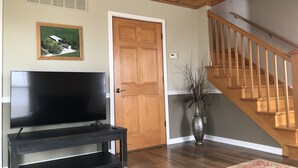 Een woonkamer met een flatscreen televisie op een donker houten dressoir, een houten deur, een trap met houten leuningen en een vaas met takken op de vloer.