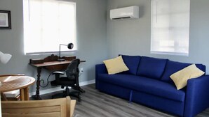 A room with a blue sofa, two windows with blinds, a desk with a chair, and an air conditioning unit.
