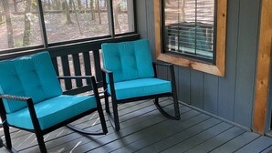 Een veranda met twee turquoise stoelen en een houten bank, grote ramen met zonwering, en een houten muur met een vogelhuisje en een wandlamp.