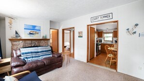 A living room with a brown leather couch, a small wooden table, and a stone fireplace. There is a kitchen visible through an open doorway. - Quaint Cabin: Beach Days & Bonfire Nights – The Ultimate Summer Escape! No Central A/C (Oscoda)