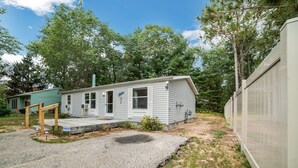 A single-story mobile home with a porch and a small deck, surrounded by trees and a gravel driveway. - Quaint Cabin: Beach Days & Bonfire Nights – The Ultimate Summer Escape! No Central A/C (Oscoda)