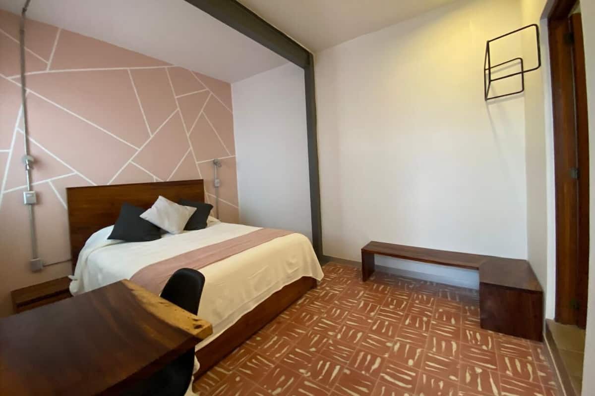 Quarto de hotel com cama, mesa de cabeceira e um banquinho.