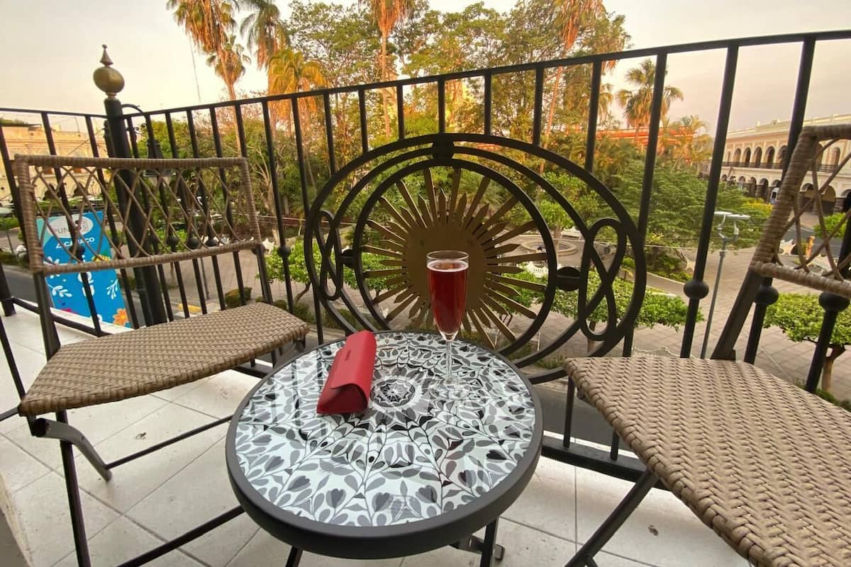Uma varanda com duas cadeiras de vime, uma mesa com um copo decorativo e vista para palmeiras e um edifício ao fundo.