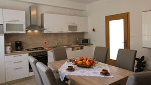 A modern kitchen with white cabinets, a gas stove, a microwave, and a dining table set with fruit and cups. - Holiday apartment mit Terrasse und Klimaanlage (Šilo)