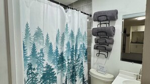 Een badkamer met een douchegordijn met bosmotief, een toilet, een handdoekhouder met opgevouwen handdoeken, en een wastafel met een spiegel erboven.