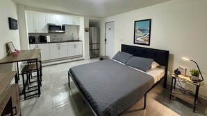Een compact studio appartement met kitchenette, een bed met hoofdeinde, een klein tafeltje en een nachtkastje met een plant.