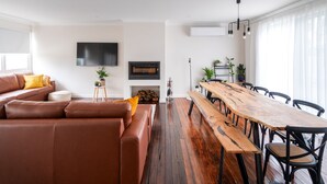 A modern living room with a brown leather sofa, a wooden dining table with chairs, a flat-screen TV mounted on the wall, and a fireplace.