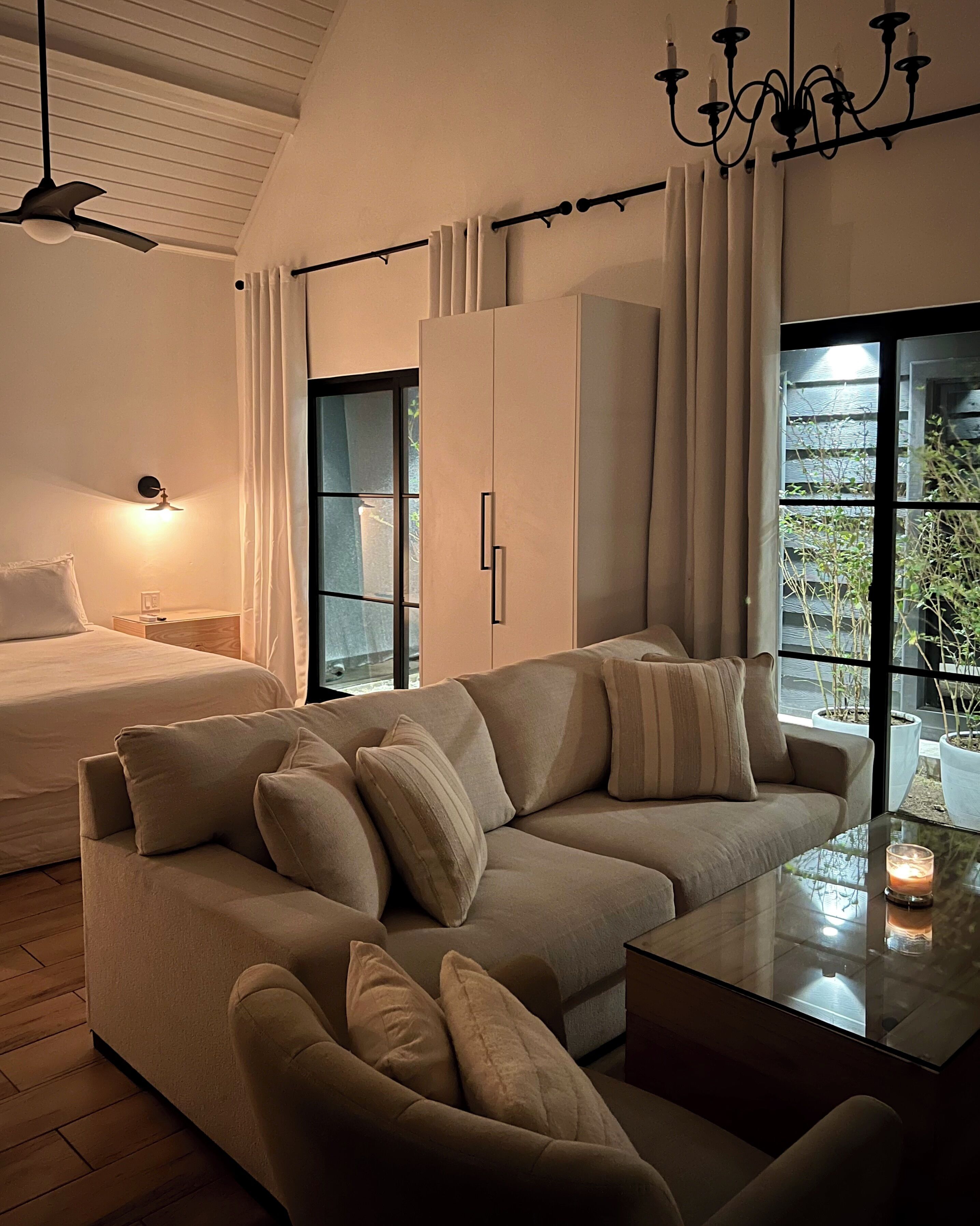 A modern bedroom with a large bed, a sofa, and a glass coffee table. There is a window with curtains and a view of greenery outside.