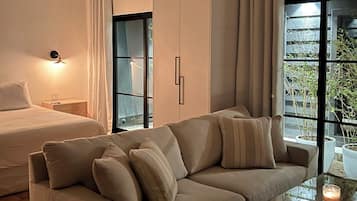 A modern bedroom with a large bed, a sofa, and a glass coffee table. There is a window with curtains and a view of greenery outside.