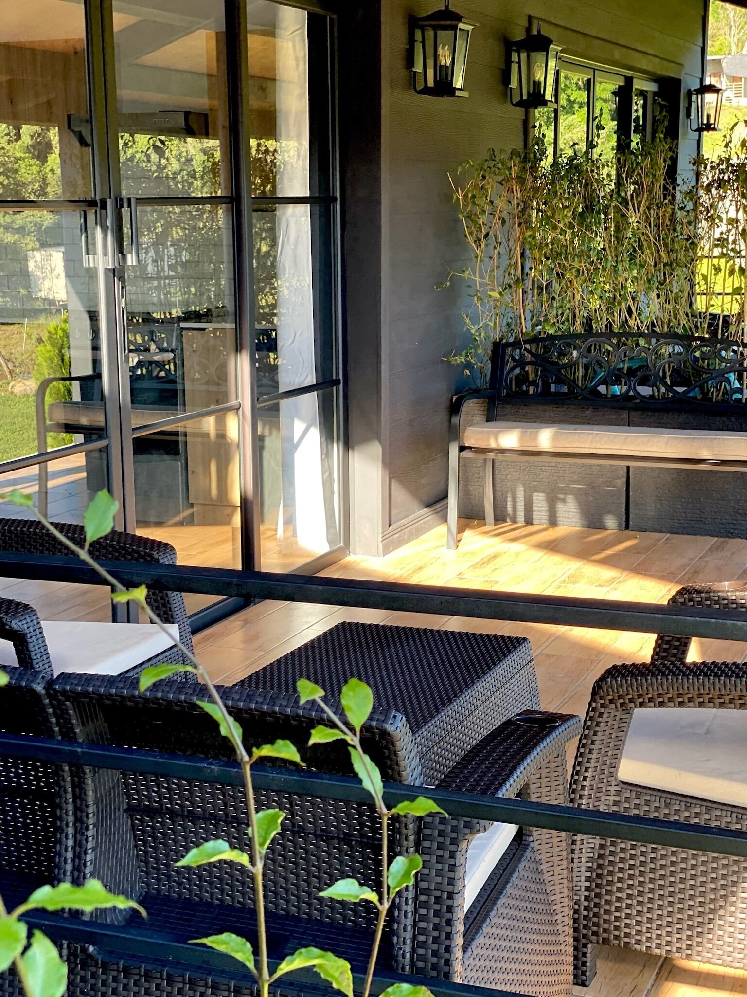 An outdoor seating area with wicker furniture and a table, surrounded by glass doors and walls, with a view of greenery outside.