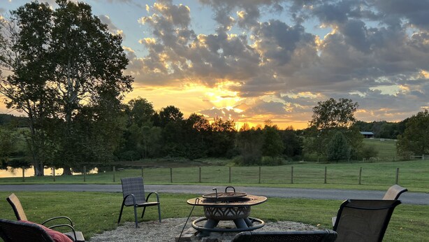 Un espace extérieur aménagé avec des chaises en osier et un foyer, offrant une vue sur un champ verdoyant et un lac, le tout baigné par les couleurs d’un ciel au coucher du soleil.