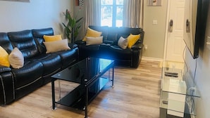 Una sala de estar con dos sofás de cuero negro, una mesa de centro de vidrio y un televisor en la pared derecha.