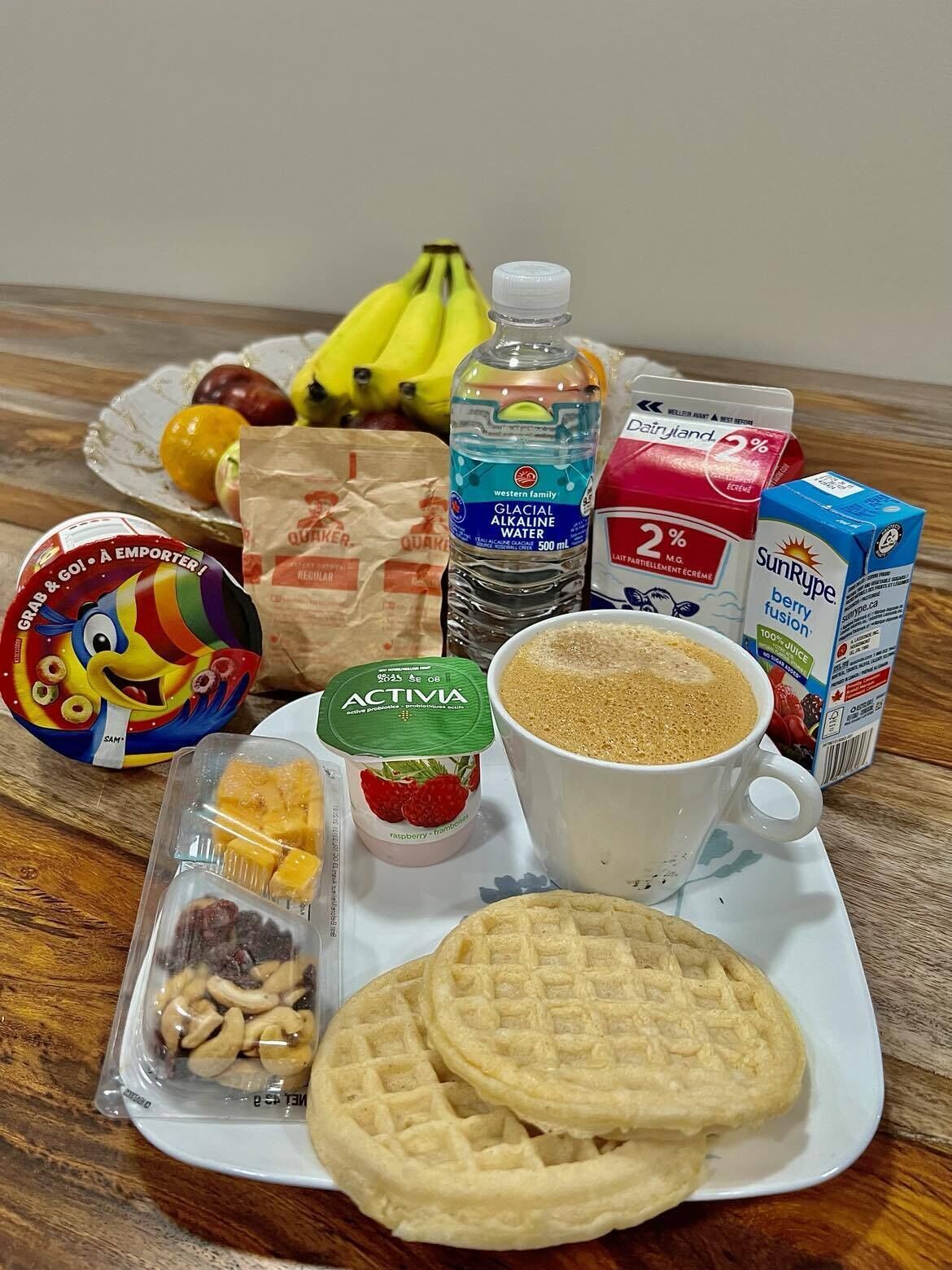 A tray with waffles, a cup of coffee, a bottle of water, a carton of milk, a bag of snacks, a jar of yogurt, a pack of cookies, and a bag of fruit.