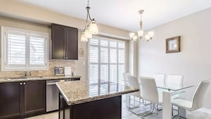 A modern kitchen with dark cabinets, a granite countertop, and a dining area with a glass table and white chairs. - Spacious and Comfortable House near Squireband (Mississauga)