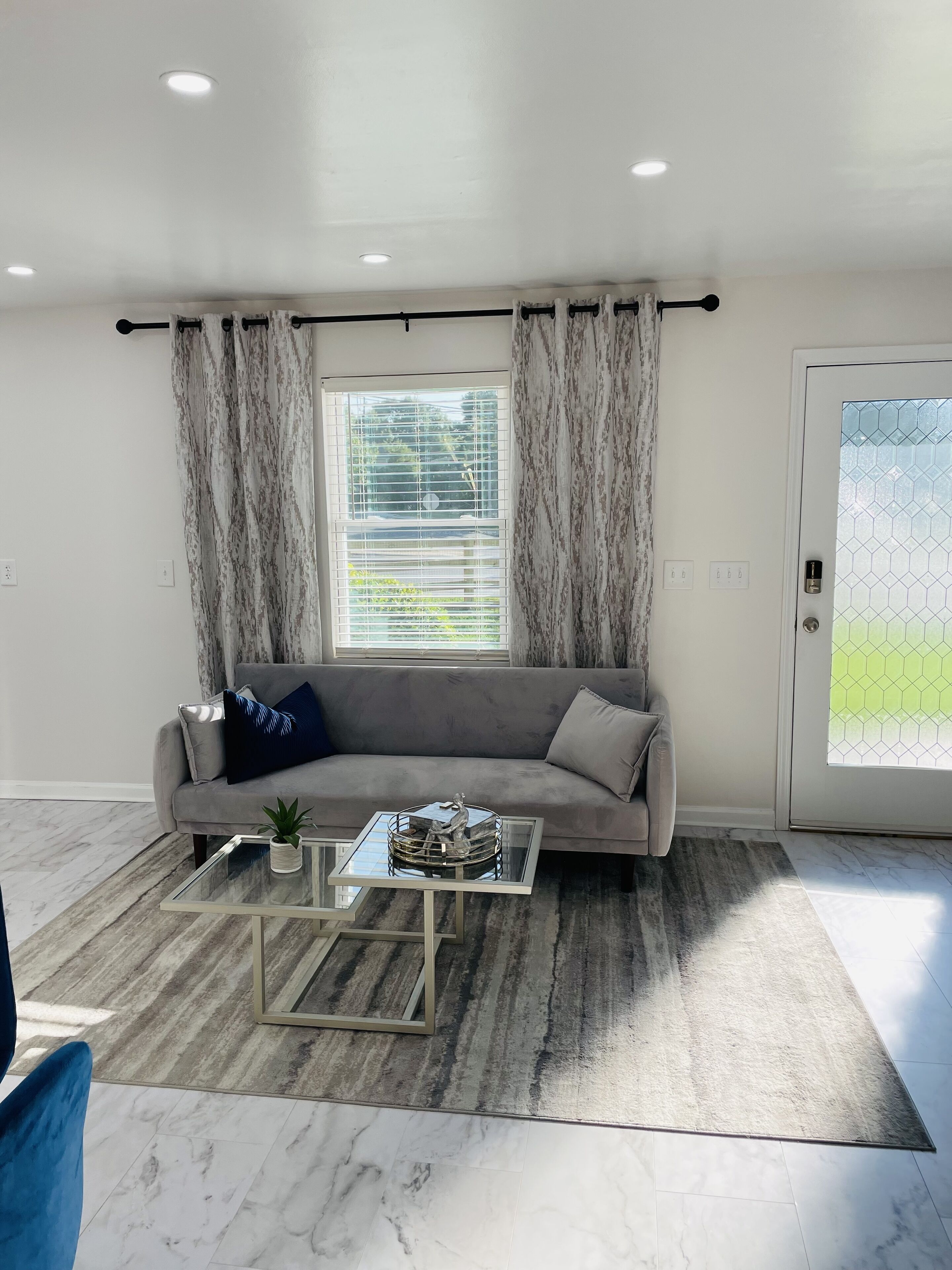 Ein modernes Wohnzimmer mit einem grauen Sofa, einem Glastisch und Blick durch ein Fenster mit Jalousien.