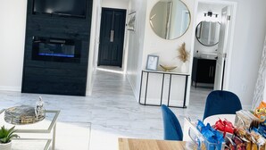 Una sala moderna con un televisor de pantalla plana, una chimenea, una zona de comedor con una mesa de madera y una cocina visible a través de un vano abierto.