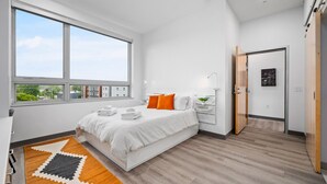 Et moderne soveværelse med et stort vindue, en seng med hvide sengetæpper og orange puder, et natbord med en lampe, og udsigt til bygninger udefra.