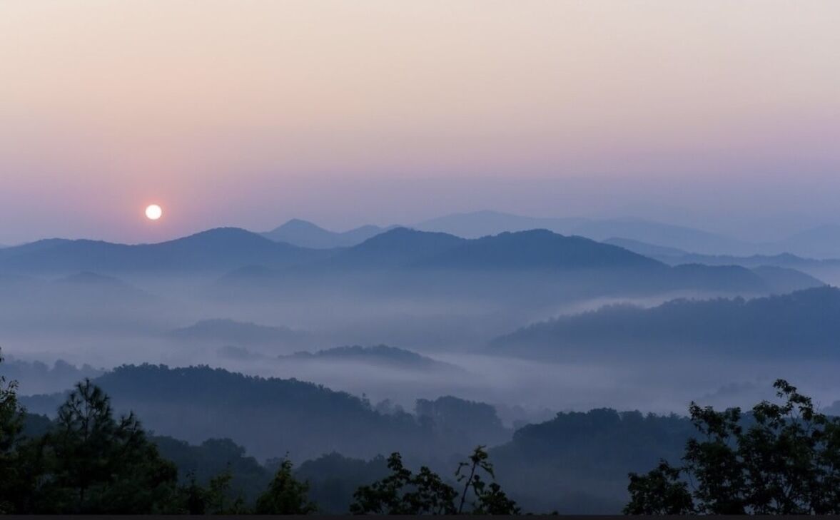 A mountainous landscape with a sunrise or sunset, featuring rolling hills and a misty atmosphere.