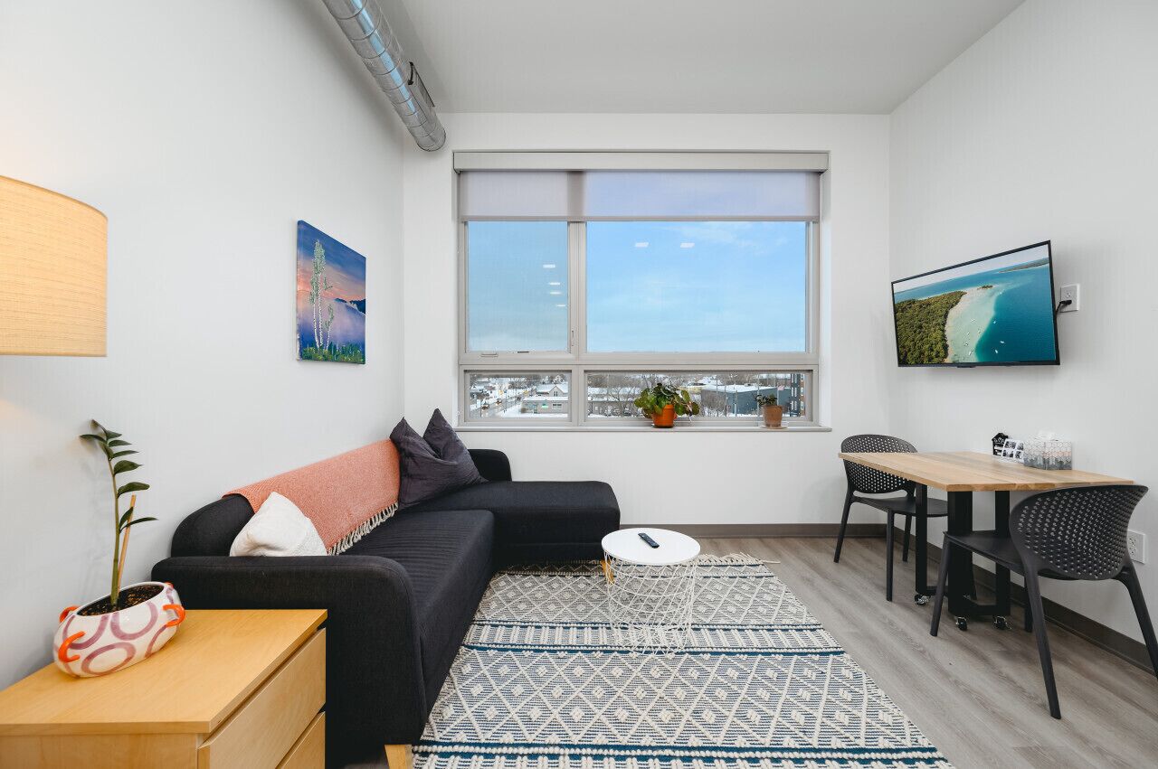 Uma sala de estar moderna com um sofá preto, uma mesinha com uma planta, um espaço de refeições com cadeiras e uma janela grande com vista para a cidade.