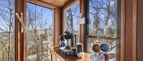 A wooden cabin interior with a coffee bar, skis leaning against the wall, and a view of a snowy landscape.